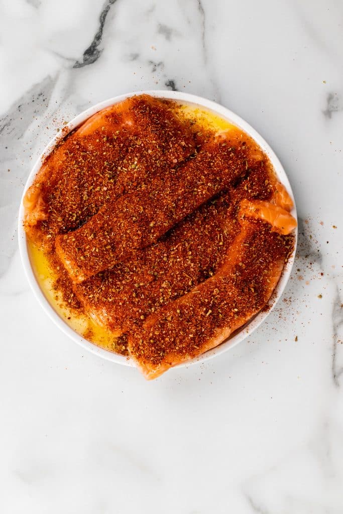 salmon filets on a white plate with melted butter topped with a thick layer of red seasoning