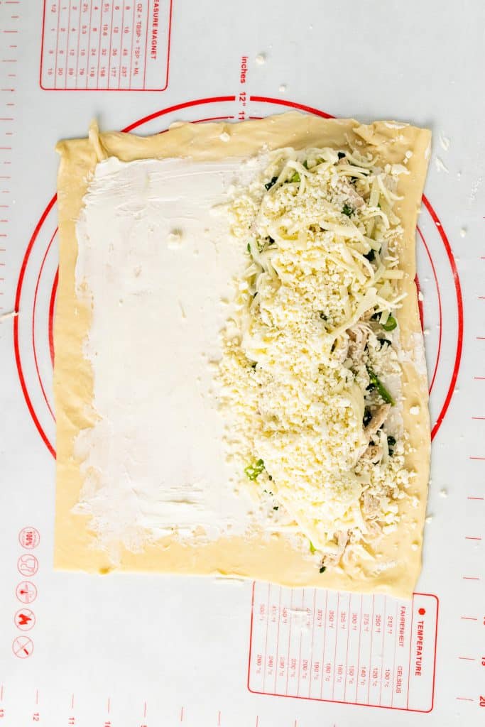 a sheet of puff pastry spread with cream cheese with spinach, chicken, and green onions arrange on one side and topped with shredded cheese
