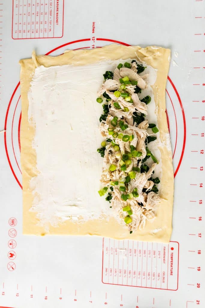 a sheet of puff pastry spread with cream cheese with spinach, chicken, and green onions arrange on one side