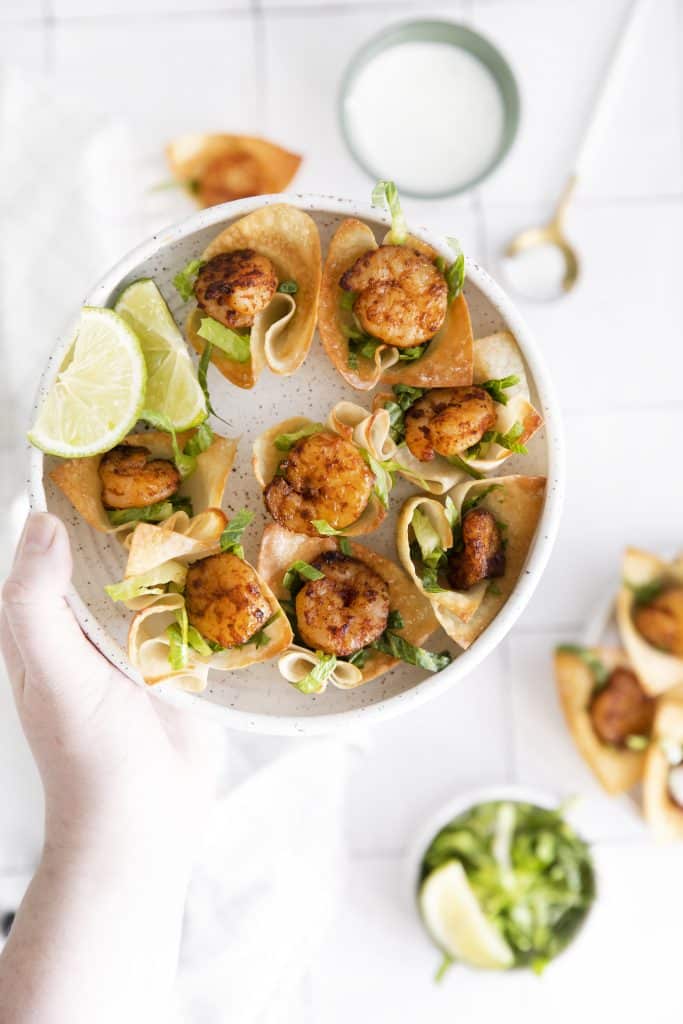 a hand holding a small white plate with shrimp taco bites and two lime wedges