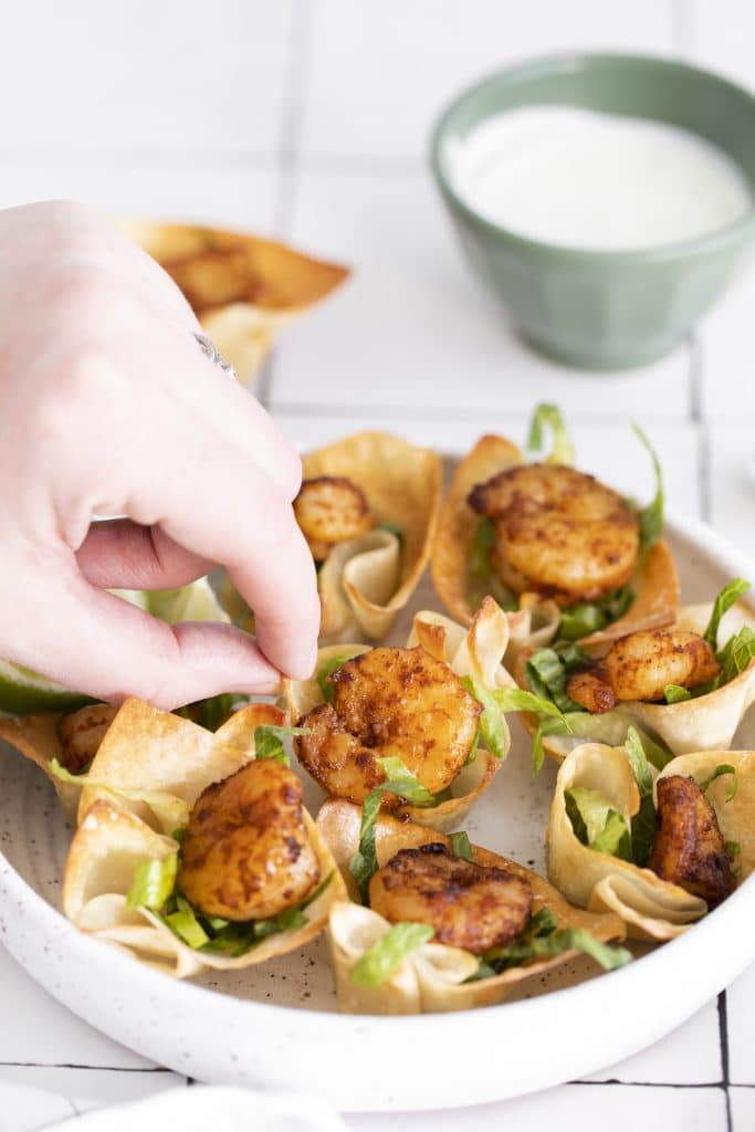 a hand lifting a wonton cup shrimp taco bite off a white plate