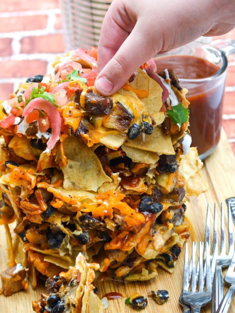 a hand shown pulling nachos from a stack of cheesy trash can nachos