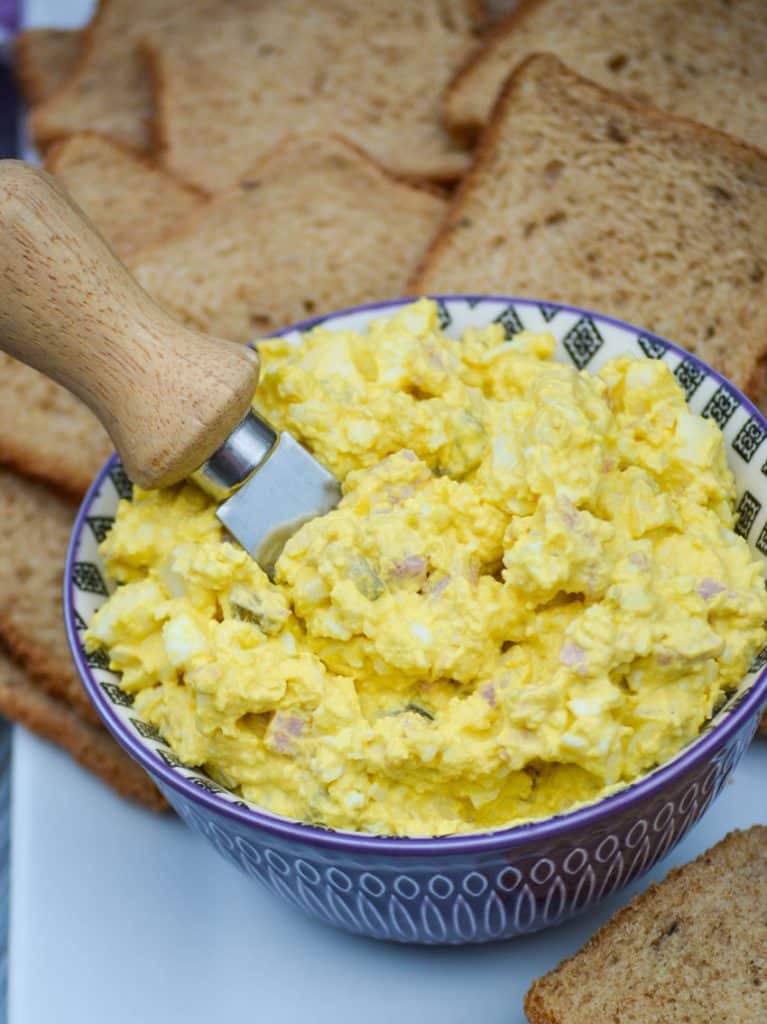 a wooden handled spreader stuck in a purple bowl filled with ham and egg salad