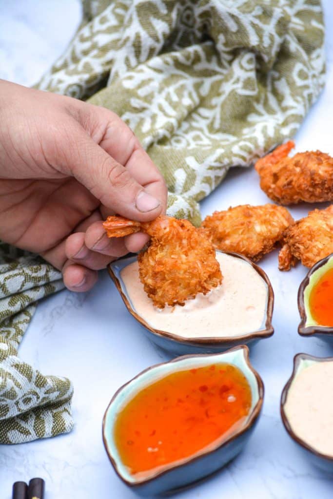 a hand dipping crispy coconut shrimp into a bowl of yumyum sauce