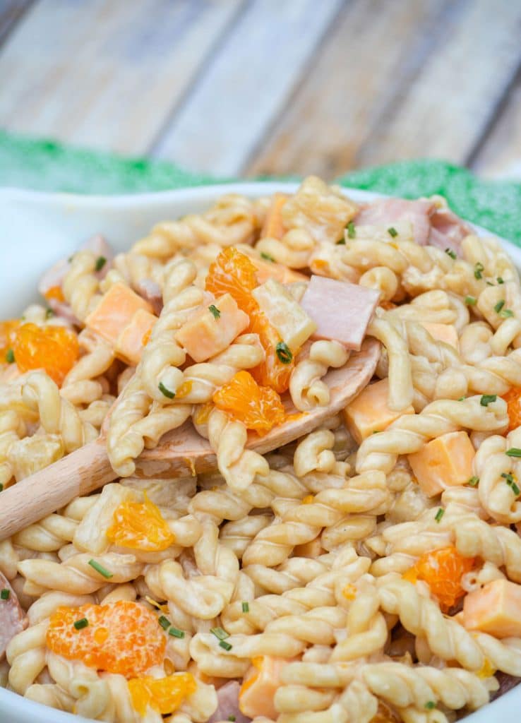 a wooden spoon digging into a bowl of creamy macaroni salad with ham, pineapple, and mandarin oranges