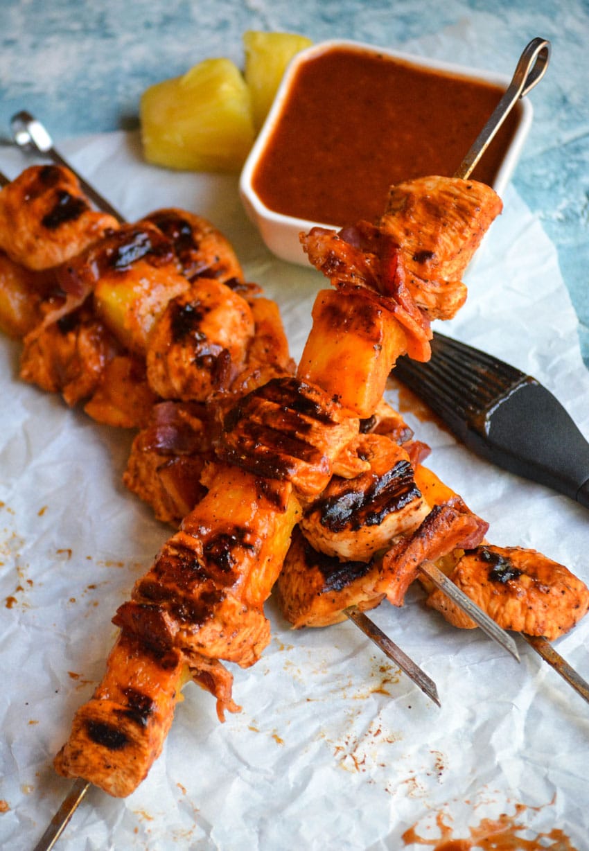 barbecue chicken kabobs with bacon and pineapple on metal skewers resting on crinkled white parchment paper