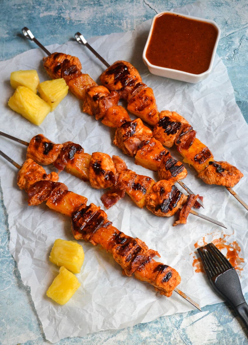 barbecue chicken kabobs with pineapple and bacon on metal skewers resting on crinkled white parchment paper