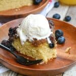 a black fork next to an ice cream topped slice of blueberry upside down cake on a wooden plate
