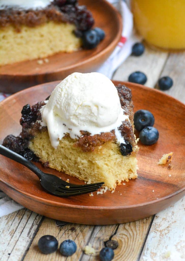 a black fork next to an ice cream topped slice of blueberry upside down cake on a wooden plate