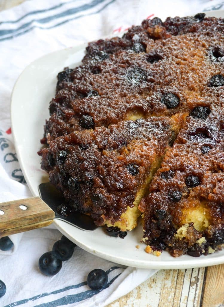 blueberry upside down skillet cake on a white platter with a slice being removed