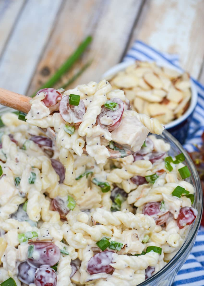 a wooden spoon lifting a scoop of creamy poppy seed chicken pasta salad out of a glass mixing bowl