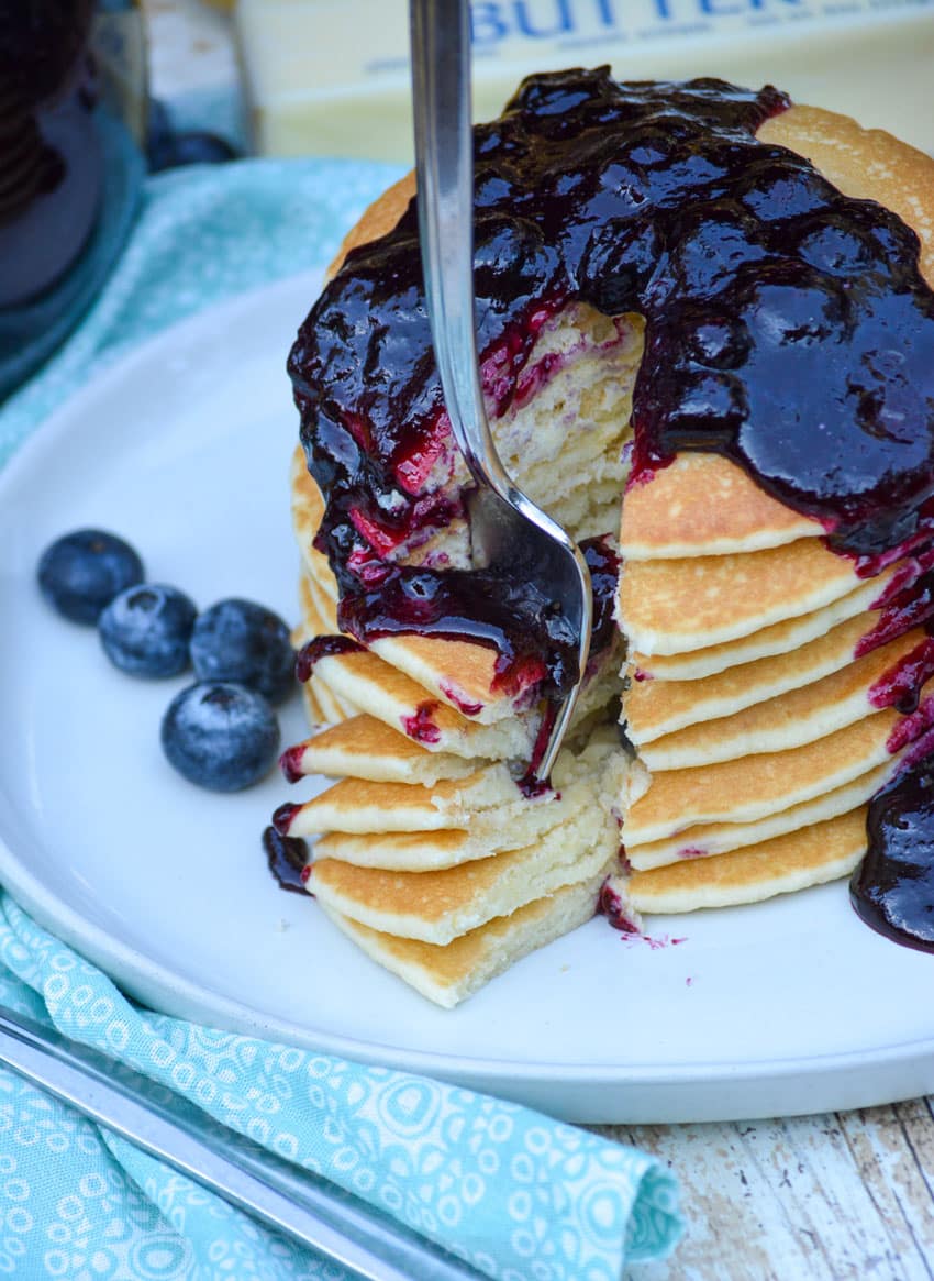 a silver fork stuck in a blueberry sauce topped stack of pancakes