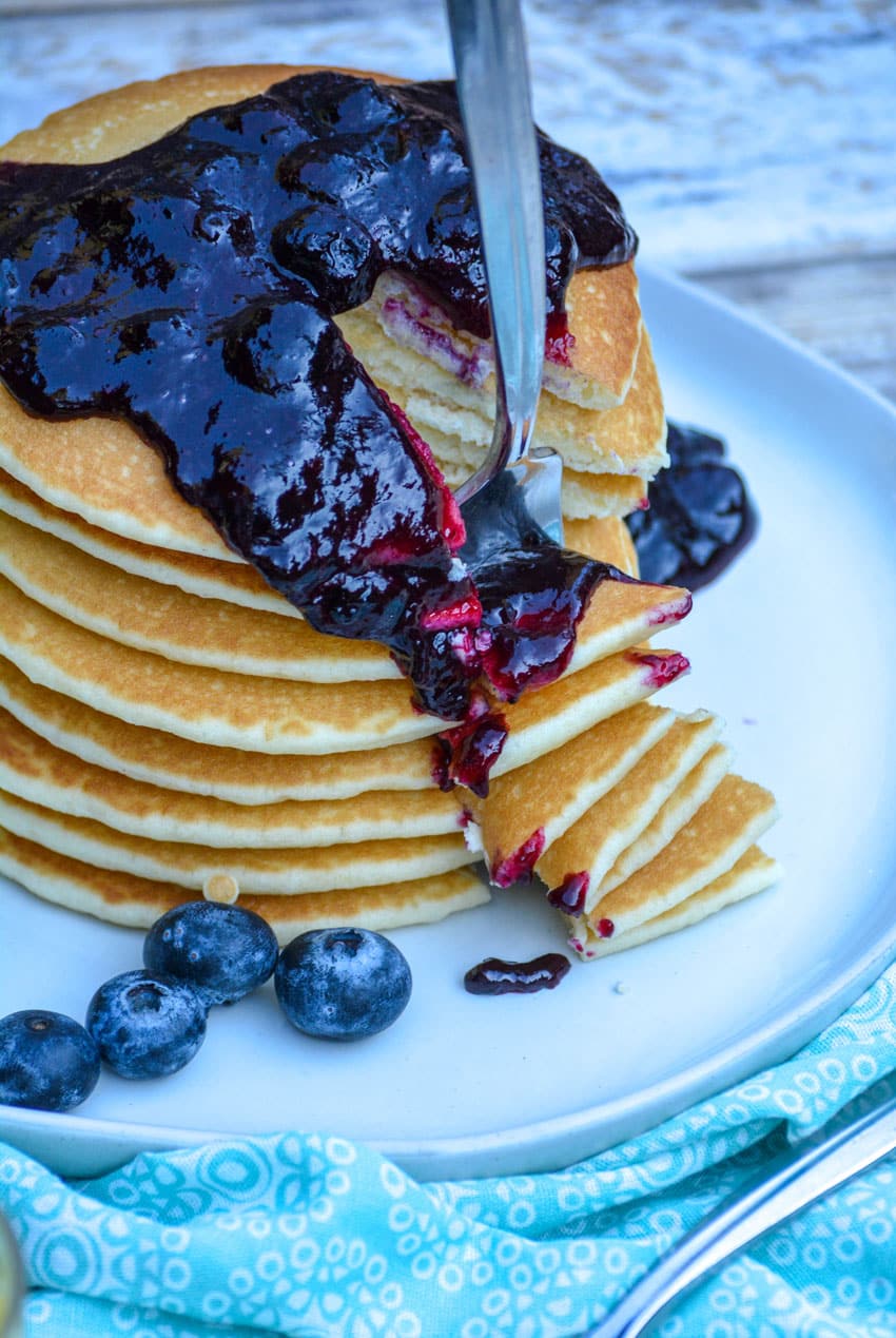 a silver fork stuck in a blueberry sauce topped stack of pnacakes