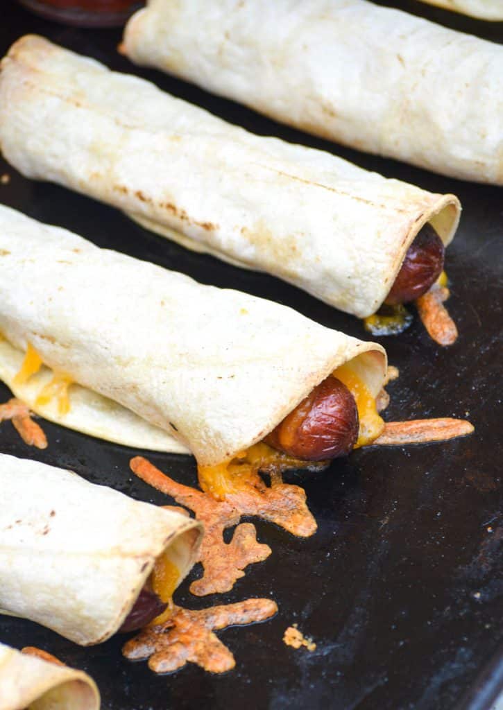 cheesy baked hot dog taquitos arranged in a row on a metal baking sheet