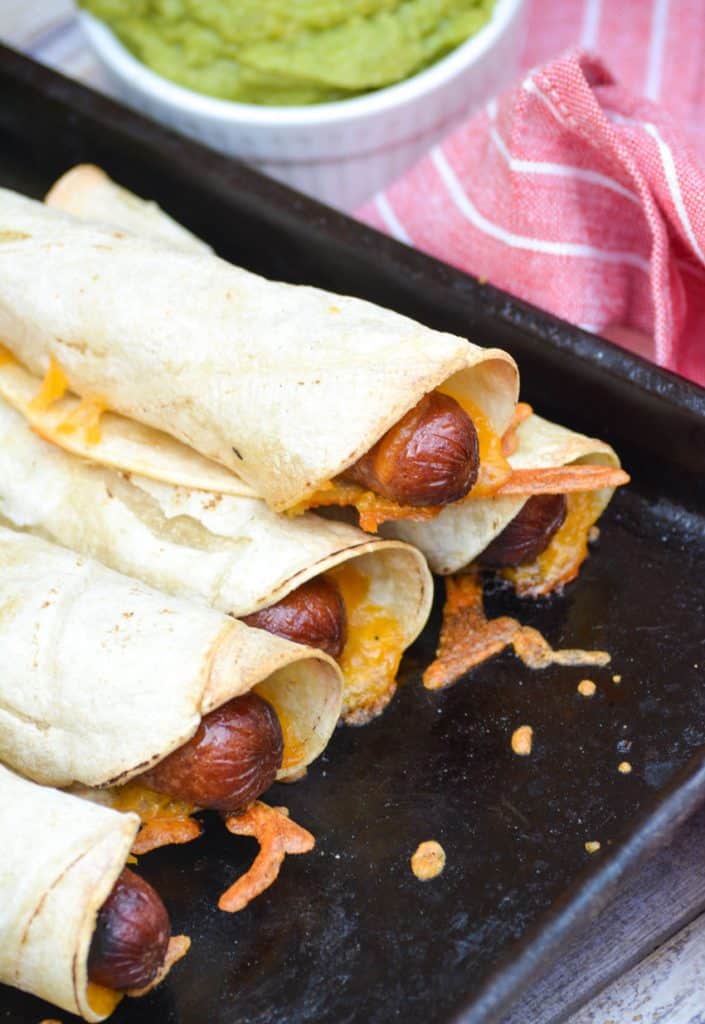 cheesy hot dog taquitos stacked on a dark baking sheet