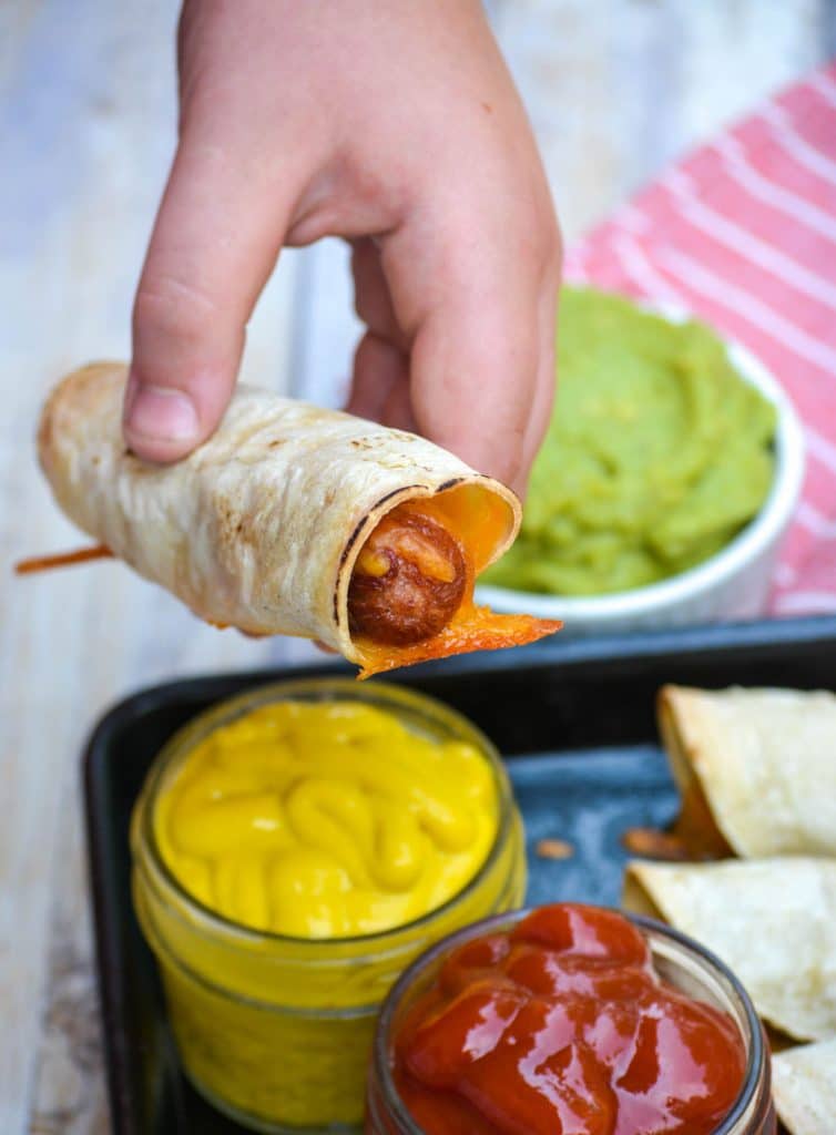 a hand holding a baked hot dog taquito over bowls of ketchup and mustard and guacamole