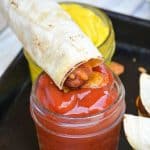 a cheesy hot dog taquito being dipped in a small glass jar of ketchup