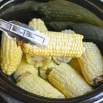 a pair of silver tongs holding a steamed piece of corn on the cob above a black crockpot