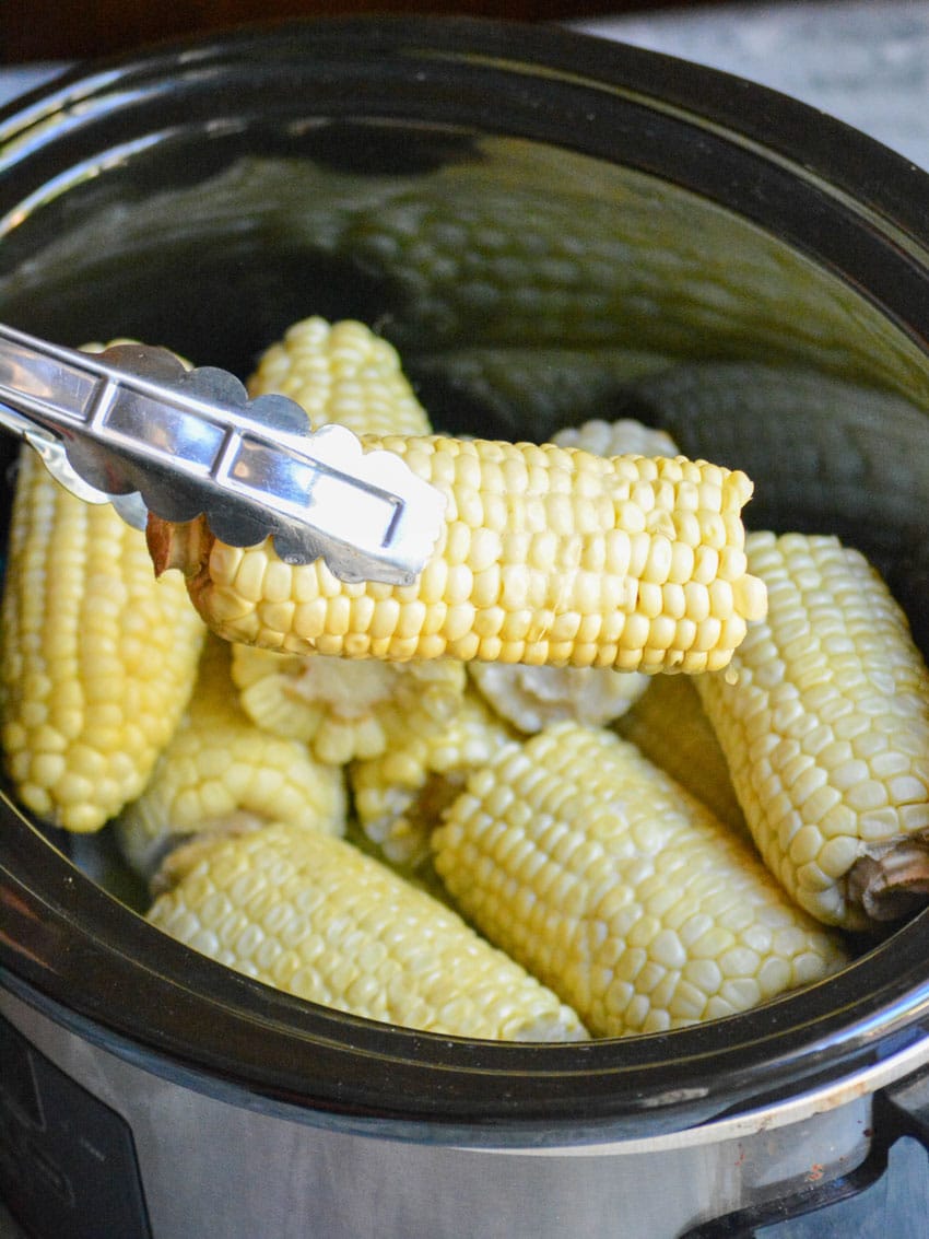 a pair of silver tongs holding a steamed piece of corn on the cob above a black crockpot