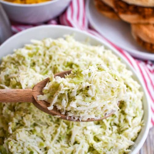 a wooden spoon holding a scoop of creamy coleslaw above a white bowl