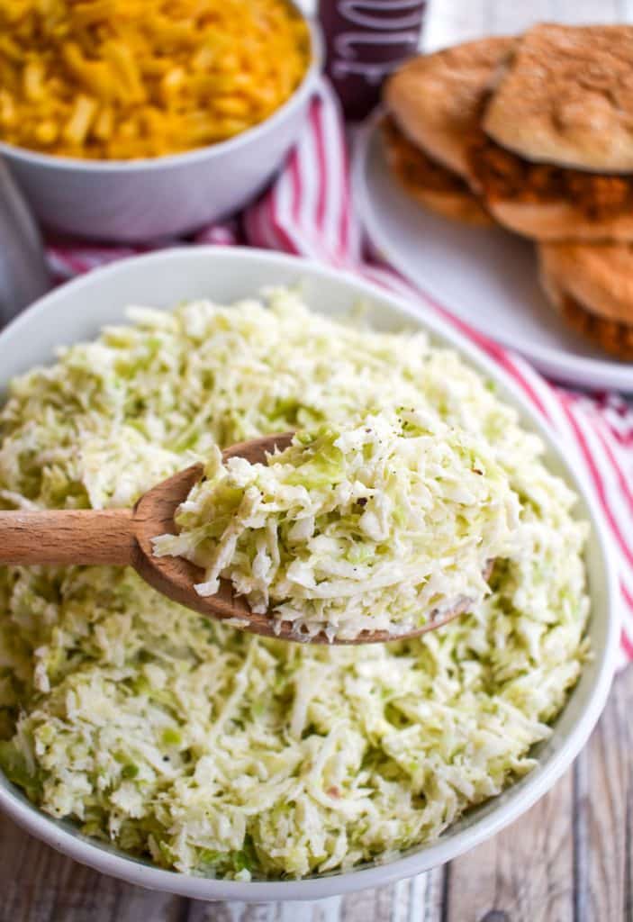 a wooden spoon holding a scoop of creamy coleslaw above a white bowl