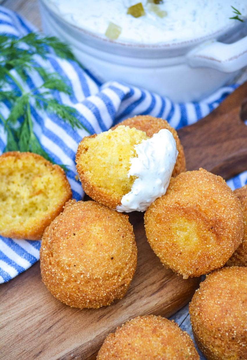 a dipped hushpuppy resting on a pile of golden brown hushpuppies on a wooden cutting board