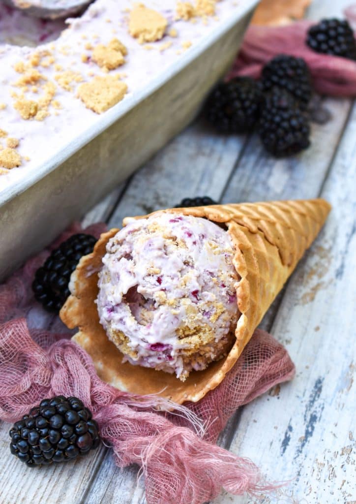 a waffle cone filled with scoops of no churn blackberry cheese cake ice cream resting on its side on a wooden table