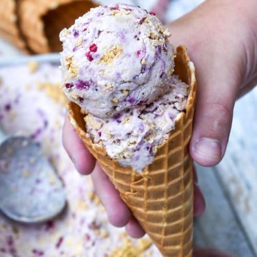 a hand holding up a waffle cone filled with scoops of no churn blackberry cheesecake ice cream