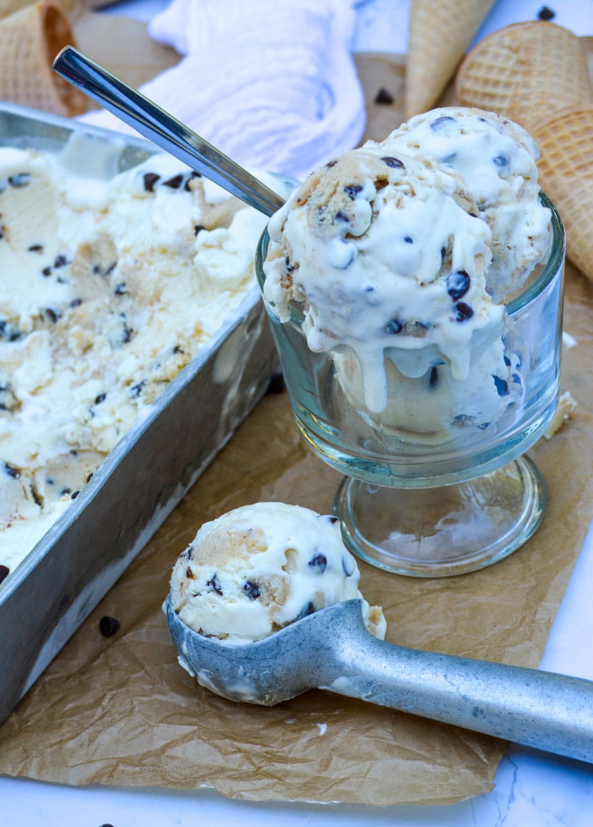 an ice cream scoop holding a scoop of cookie dough ice cream sitting on a sheet of brown parchment paper in front of a glass bowl filled with melting ice cream