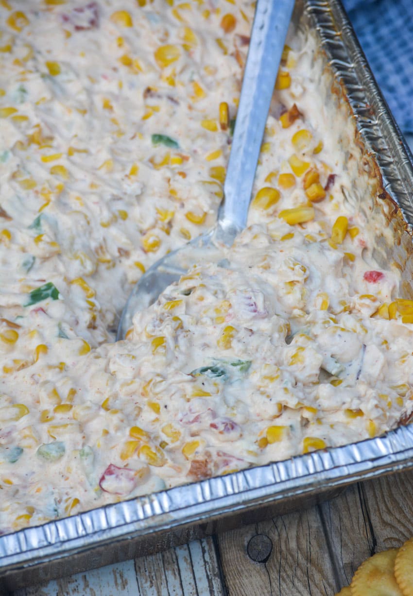 a silver serving spoon stuck into smoked creamy corn dip in a disposable aluminum pan