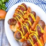 mustard and ketchup drizzled air fryer beer brats arranged in a row on a white serving platter
