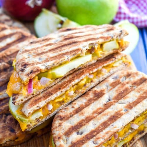 slices of apple cheddar paninis on a wooden cutting board with a red onion and green apple on the side