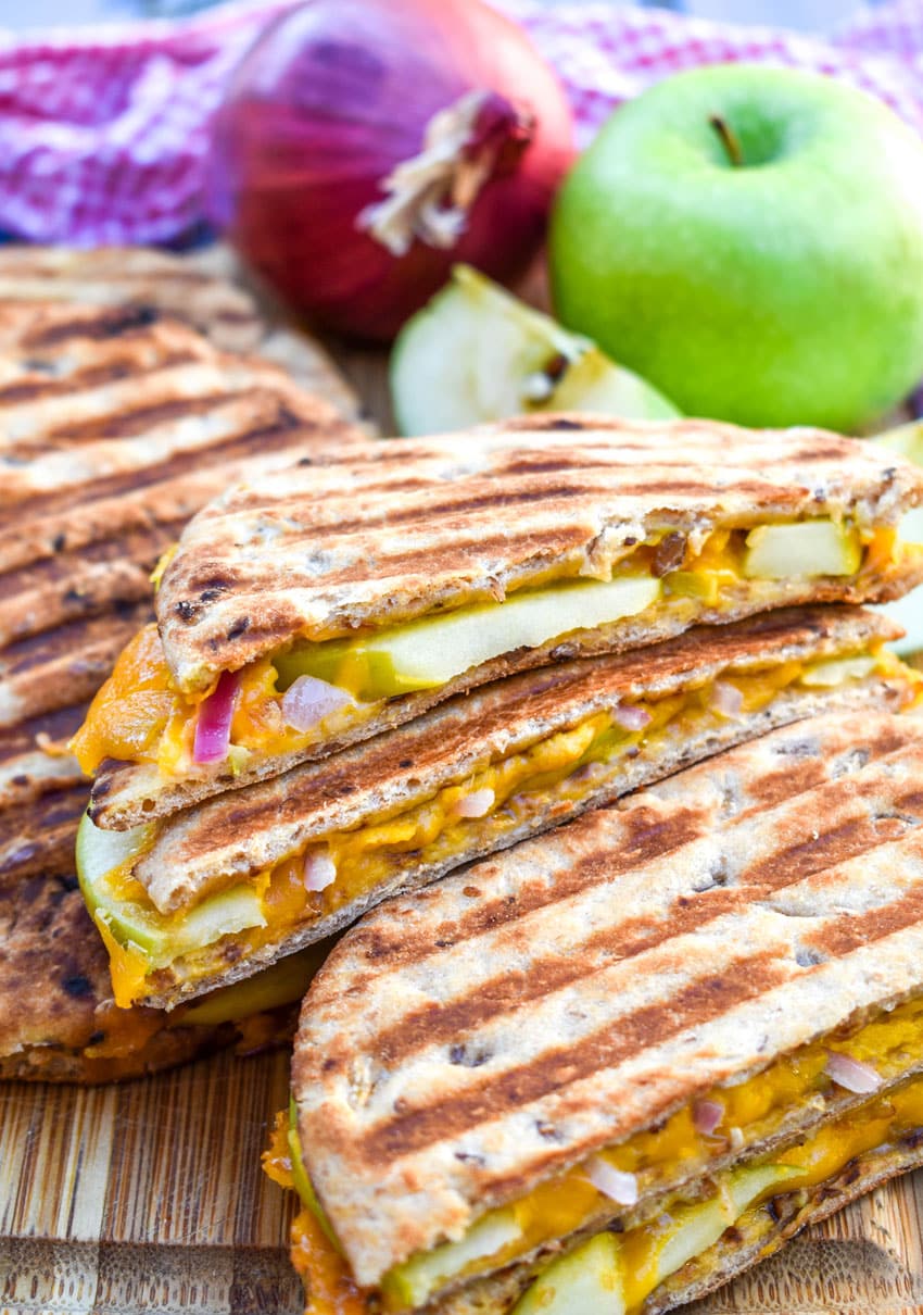 slices of apple cheddar paninis on a wooden cutting board with a red onion and green apple on the side