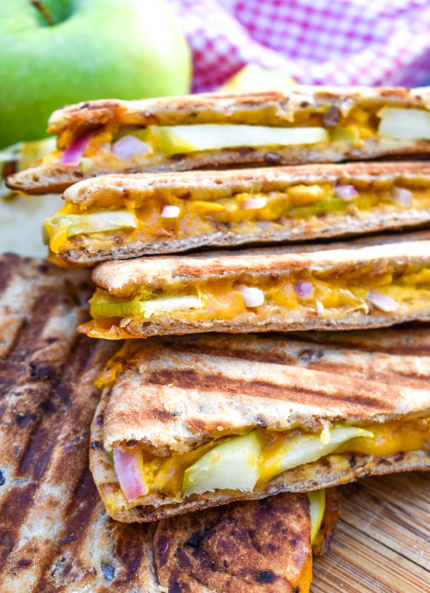 slices of apple cheddar paninis on a wooden cutting board with a red onion and green apple on the side