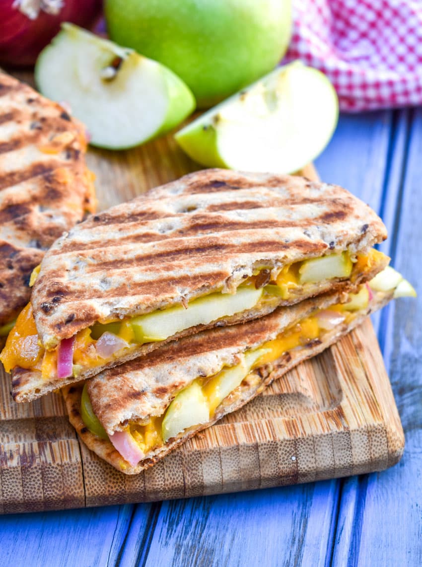 slices of apple cheddar paninis on a wooden cutting board with a red onion and green apple on the side