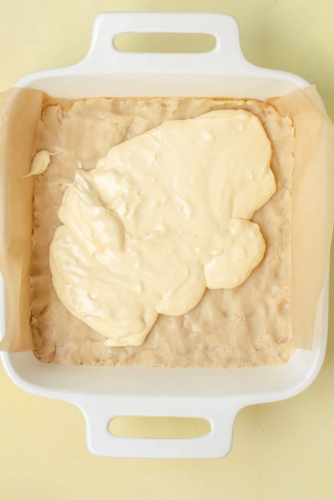 creamy cheesecake mixture poured onto a layer of sugar cookie dough in a parchment paper lined baking dish