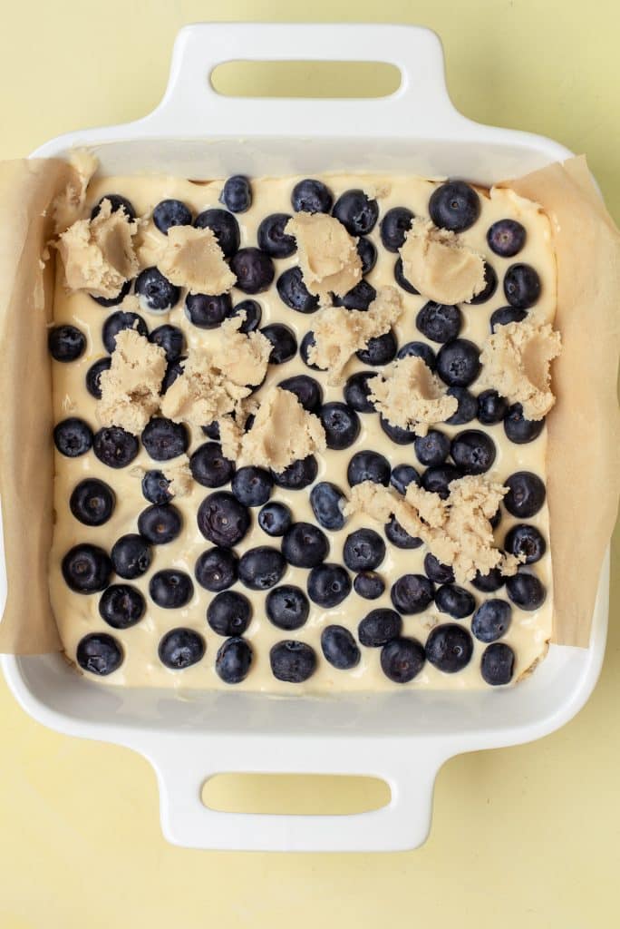 unbaked blueberry cheesecake sugar cookie bar layers shown in a parchment paper lined baking dish
