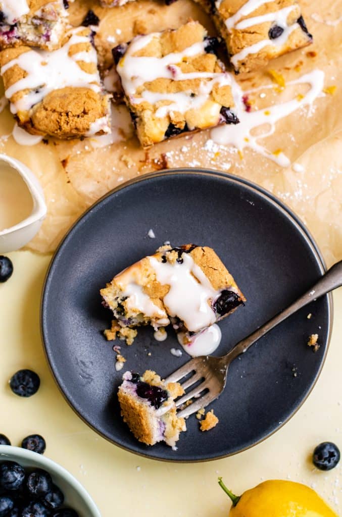 a black fork with a piece of blueberry cream cheese topped sugar cookie bars on a dark blue dessert plate