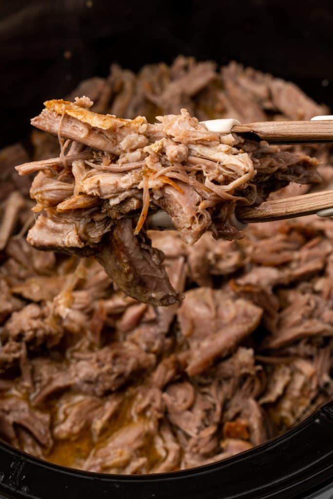 a pair of tongs holding up a serving of crockpot pulled pork over a black crockpot