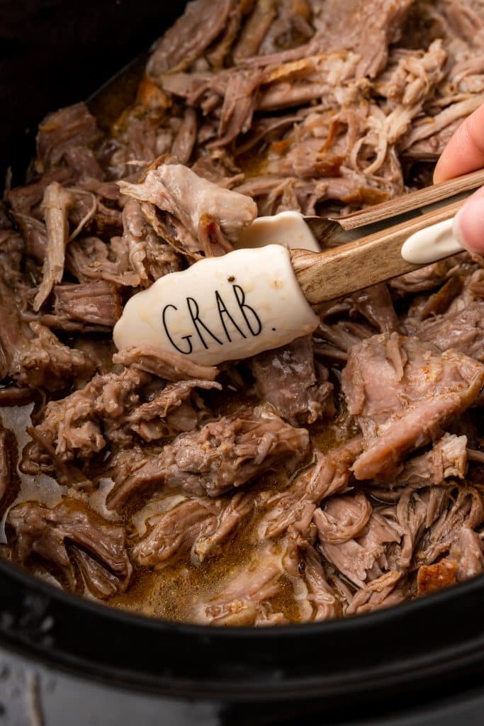a pair of tongs in a slow cooker filled with pulled pork