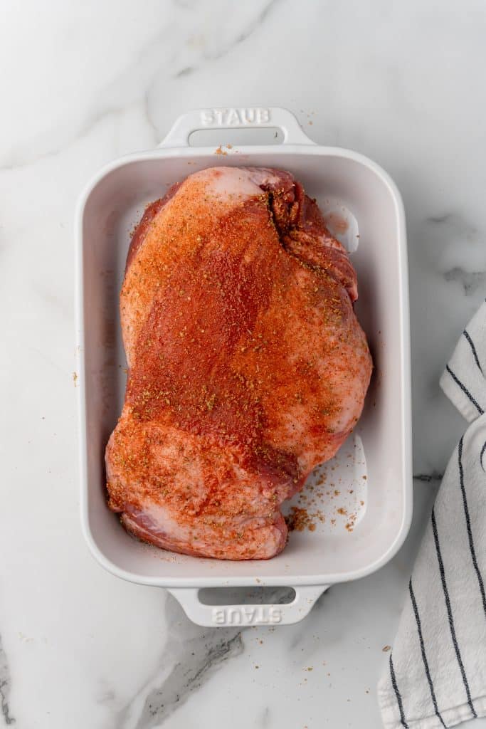 a rubbed down raw pork shoulder  sitting in a white casserole dish