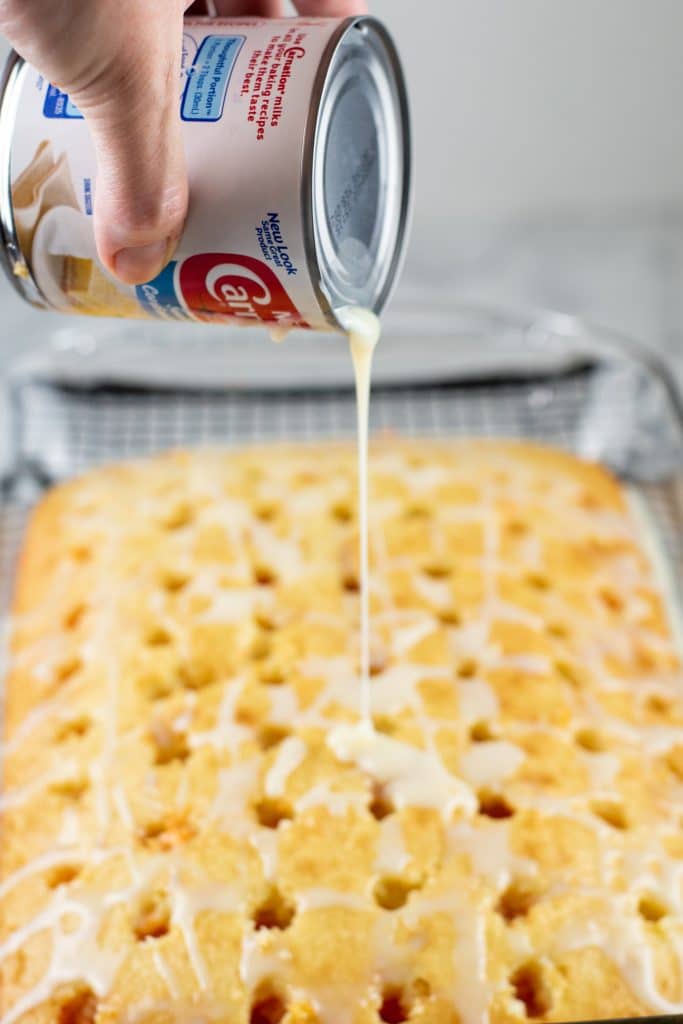 a can of condensed milk being poured over a cake with holes poked all over it