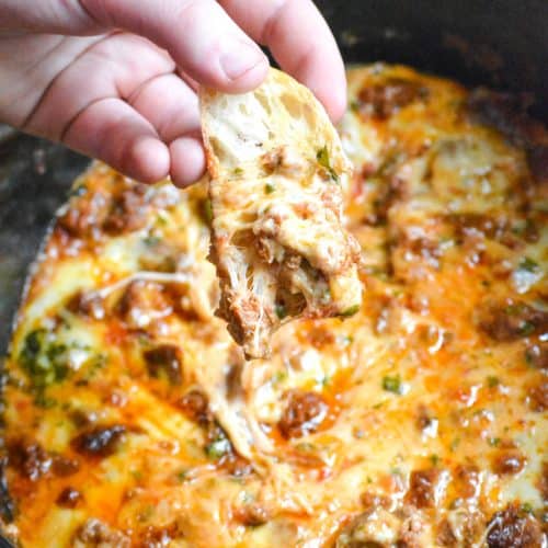 a hand holding a baguette slice topped with slow cooker queso fundido above a corckpot filled with cheesy dip