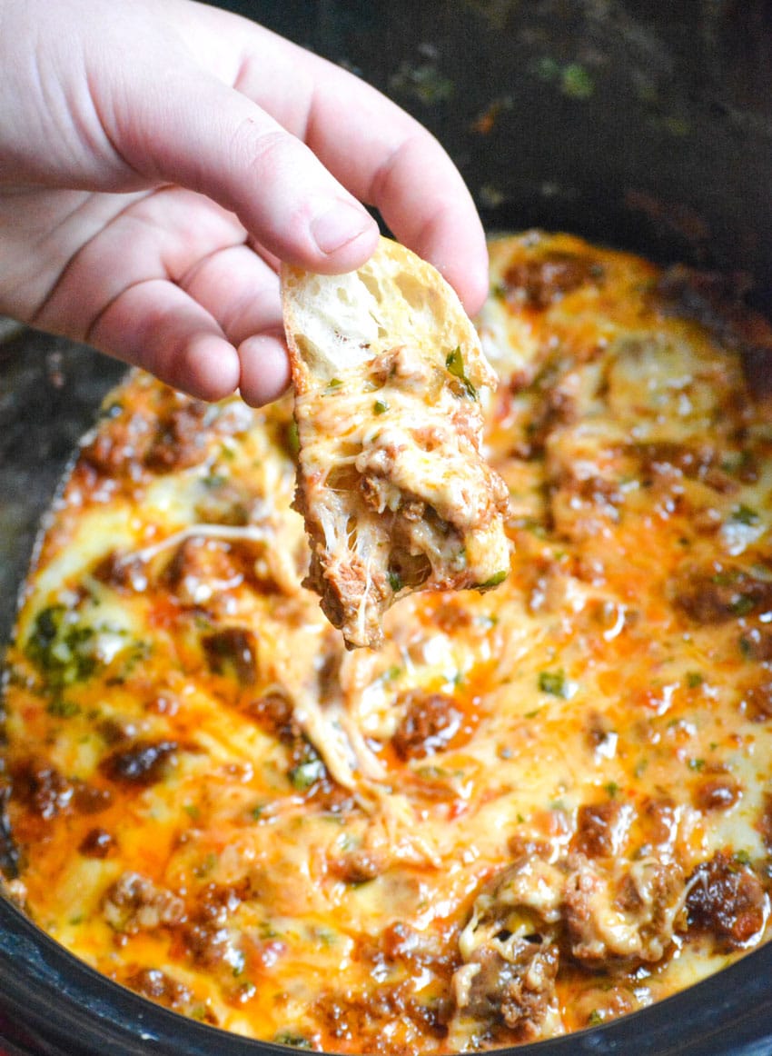 a hand holding a baguette slice topped with slow cooker queso fundido above a corckpot filled with cheesy dip