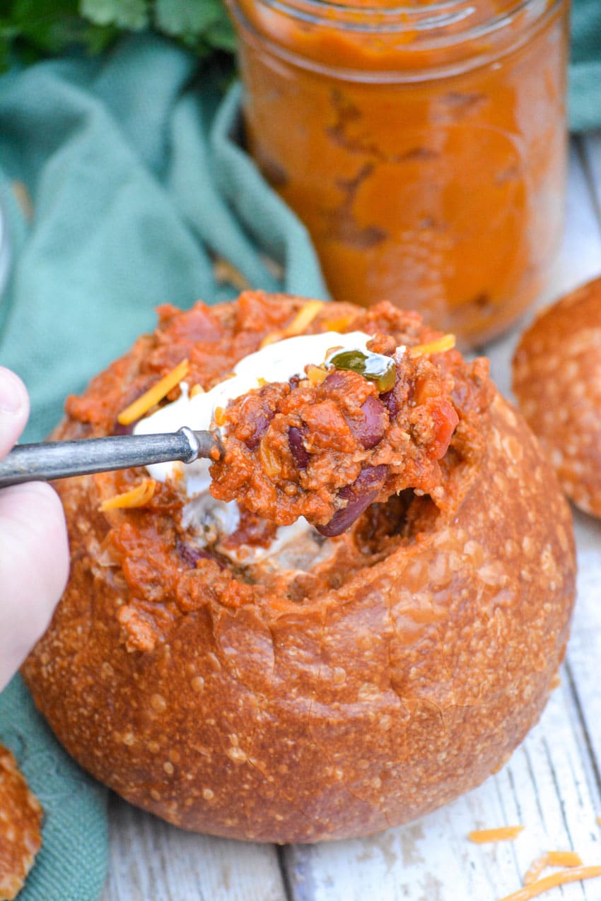 a silver spoon lifting a scoop of hearty beef & bean chili with pumpkin out of a hollowed out bread bowl