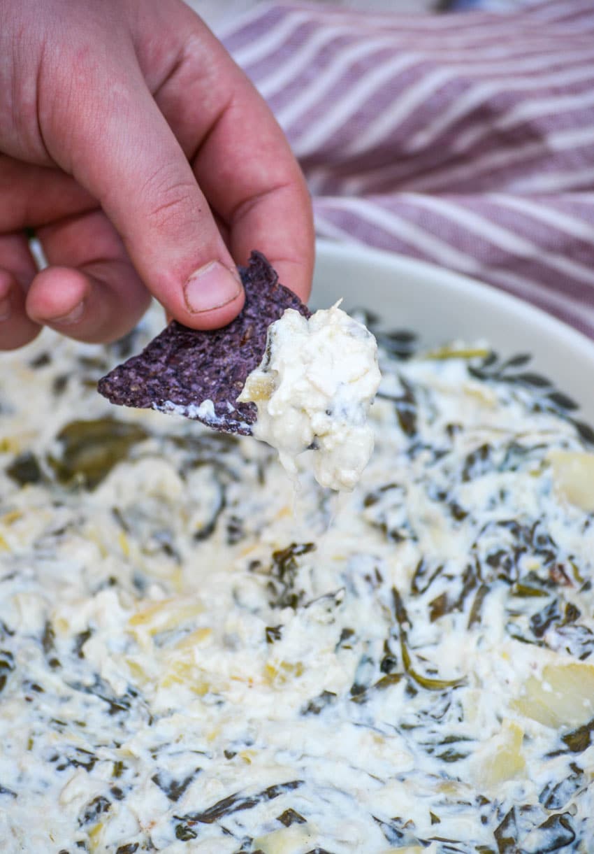 a hand holding up a blue tortilla chip topped with crockpot spinach and artichoke dip