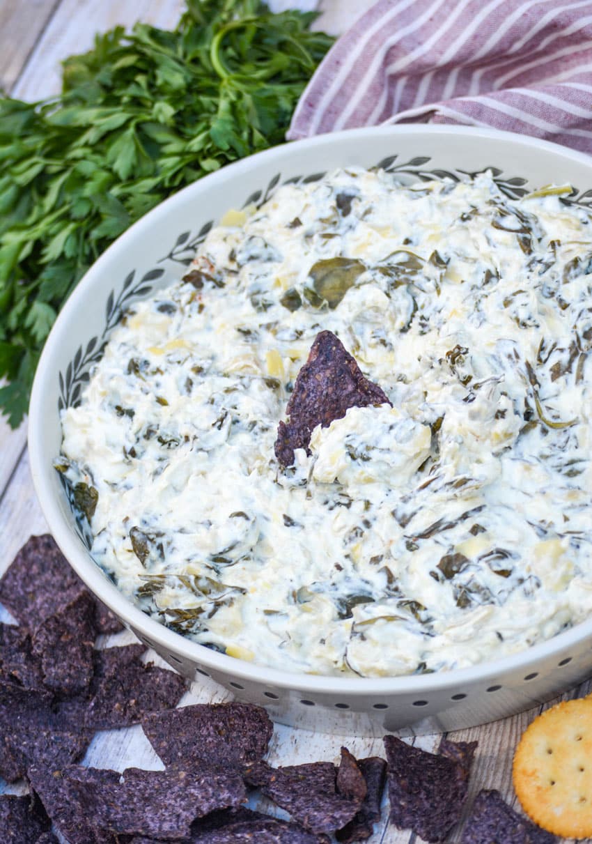 a blue corn tortilla stuck in the center of a white serving dish filled with slow cooker spinach artichoke dip