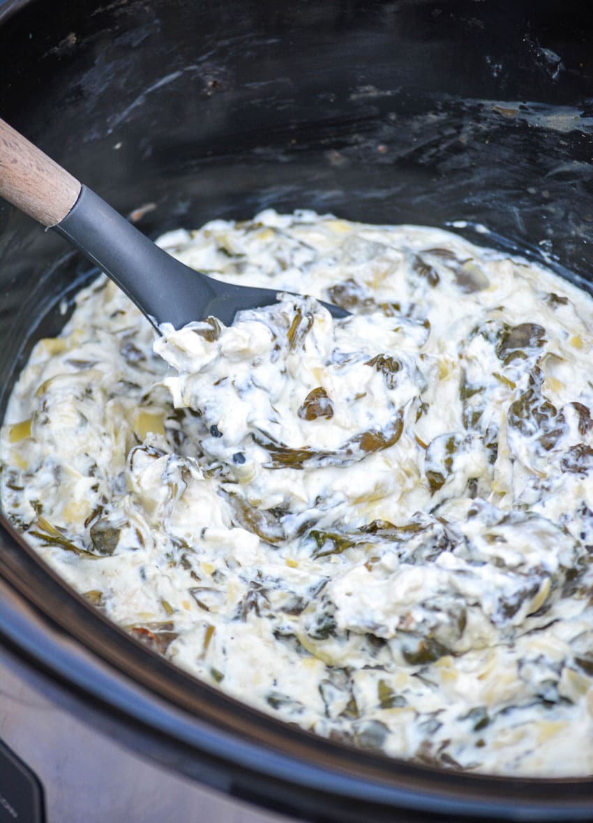 a spoon scoopinn spinach and artichoke dip out of a black slow cooker