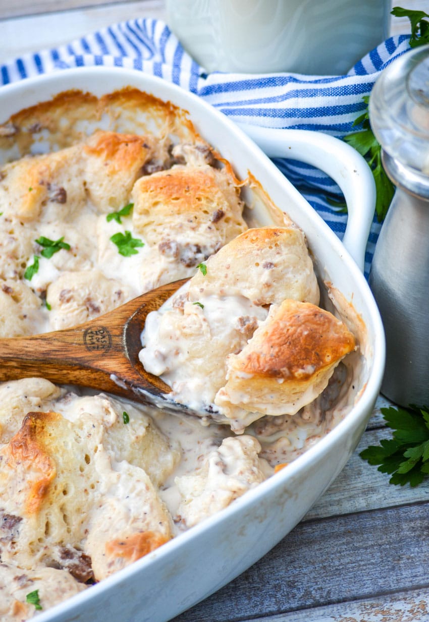 a wooden spoon in a sausage gravy and biscuits casserole in a white dish