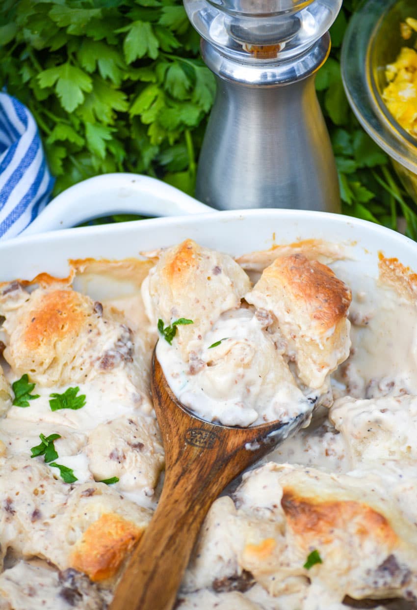 a wooden spoon in a sausage gravy and biscuits casserole in a white dish
